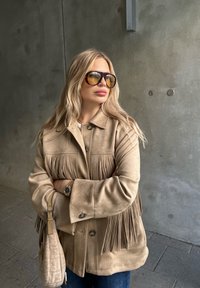 Bež jakna iz nobljenega usnja s frufru detajli, dvema gumboma in zareznim ovratnikom. Obučen over belo srajco in kombiniran s temnimi kavbojkami ter sončnimi očali.