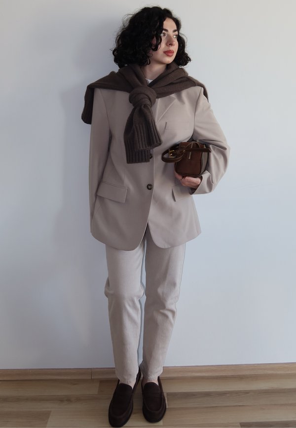 Femme portant un costume beige avec une écharpe marron foncé nouée autour des épaules, tenant un petit sac à main marron, debout sur un sol en bois.