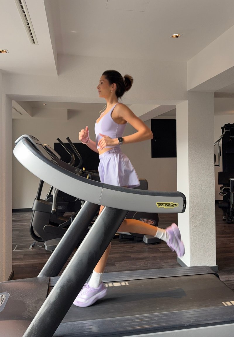 Γυναίκα με ανοιχτό μωβ αθλητικά ρούχα τρέχει σε διάδρομο Technogym σε ένα μοντέρνο, φωτεινό γυμναστήριο με ξύλινα δάπεδα.