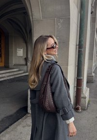 Femme aux longs cheveux blonds portant des lunettes de soleil et un manteau gris, portant un sac en cuir tressé marron, debout près d'une arche en pierre d'un bâtiment.