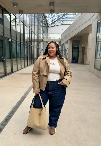 Chaqueta corta beige sobre una blusa blanca con recortes, combinada con vaqueros de tiro alto azul y un bolso beige. Zapatillas marrones completan el conjunto.