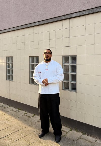 Hombre con el pelo trenzado y gafas que lleva una camisa blanca de manga larga, pantalones negros y joyas de oro, de pie junto a una pared embaldosada con pequeñas ventanas de cristal.