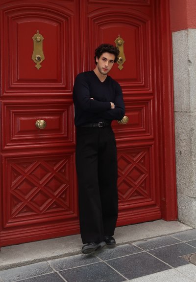 Modelo vistiendo un suéter ajustado de color azul marino oscuro y pantalones negros, de pie contra una puerta roja con un diseño de diamantes y herrajes de bronce.