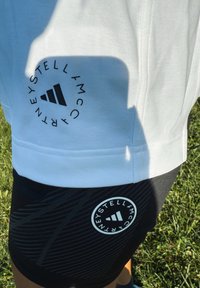 Un top alb și pantaloni scurți negri cu logo-uri circulare Stella McCartney Adidas, purtați în aer liber pe iarbă sub lumina strălucitoare a soarelui.