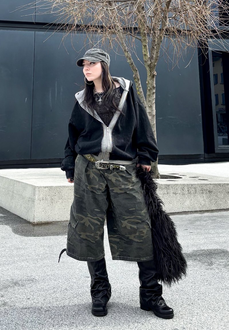 Jeune personne en short camouflage, veste zippée noire oversize, bottes noires, casquette grise, tenant un accessoire noir poilu, debout près d'un arbre nu en extérieur.