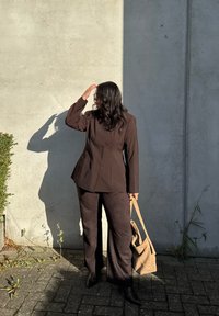 Brun skræddersyet blazer med peplum-design, matchende brune wide-leg bukser og en vævet tan håndtaske, sat mod en grå væg.