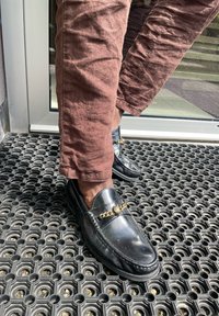 Mocasines de cuero negro con un detalle de cadena dorada, combinados con pantalones marrones texturizados, de pie sobre una alfombrilla negra con rejilla cerca de una puerta de vidrio.