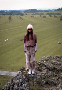 Donna con un berretto rosa, occhiali da sole e tuta marrone in piedi su un terreno roccioso che guarda un campo verde con collina lontane.