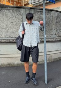 Šviesiai mėlynas raštuotas ilgas rankovės marškinėliai, juodi pritaikyti šortai, juodi elegantiški batai ir tamsi kuprinė. Stovi šalia gatvės stulpo.