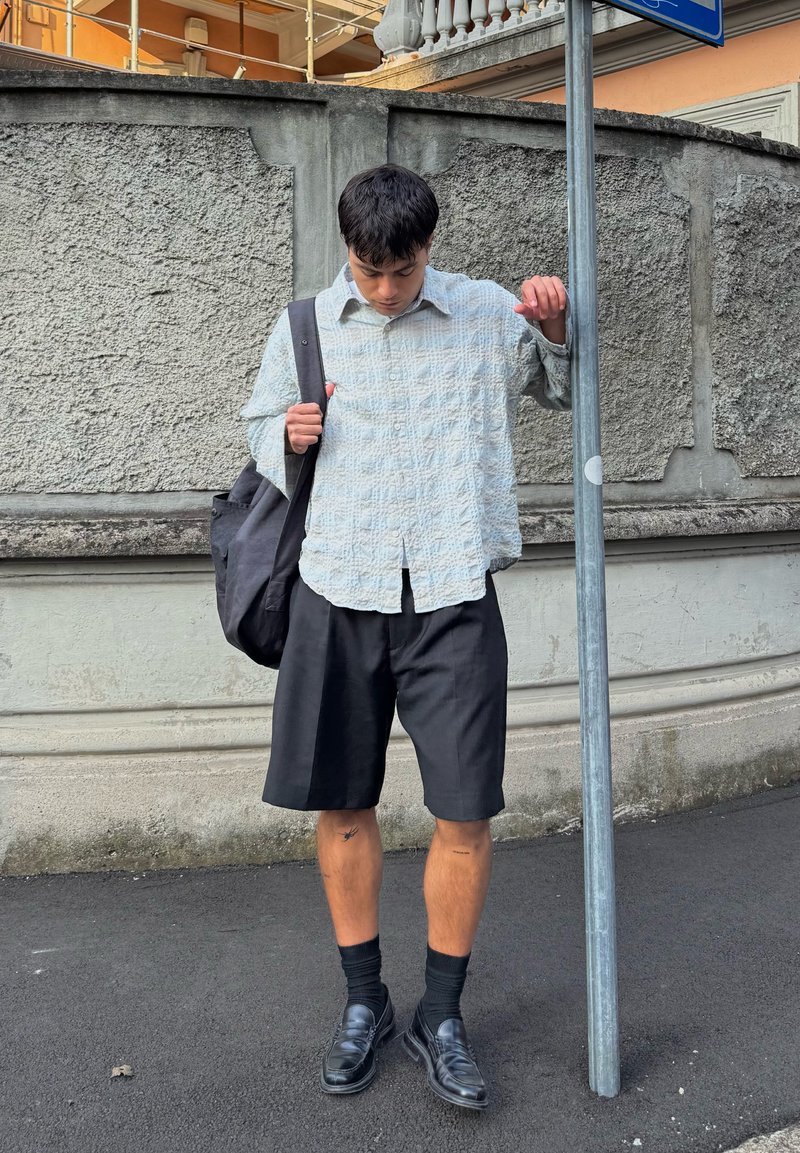 Camicia a maniche lunghe azzurro chiaro a fantasia, pantaloni corti neri su misura, scarpe eleganti nere e uno zaino scuro. In piedi accanto a un palo stradale.