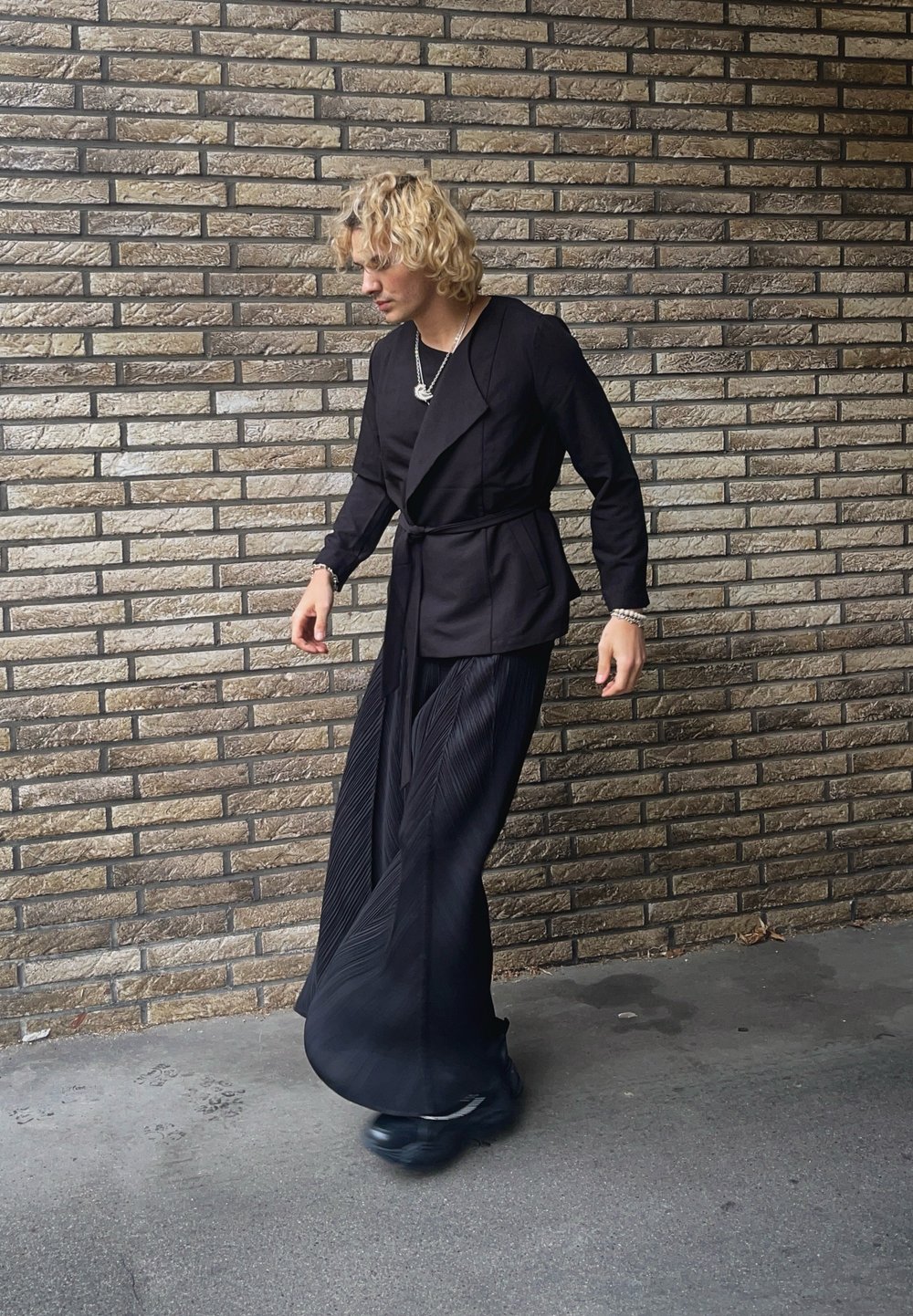 Μαύρο σακάκι με μακριά μανίκια, ζώνη στη μέση και πλισέ παντελόνι με φαρδιά πόδια. Τα αξεσουάρ περιλαμβάνουν στρωματωμένα κολιέ και μαύρα υποδήματα.