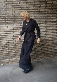 Μαύρο σακάκι με μακριά μανίκια, ζώνη στη μέση και πλισέ παντελόνι με φαρδιά πόδια. Τα αξεσουάρ περιλαμβάνουν στρωματωμένα κολιέ και μαύρα υποδήματα.