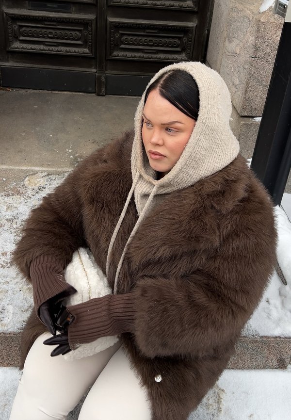 Vrouw in een bruine bontjas, beige trui met capuchon, zwarte handschoenen en witte broek zittend op besneeuwde trappen bij een donkere houten deur.