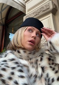 Krem- og brunspraglete fuskepelskåpe med svart filthat. Kåpen har en myk tekstur og et voluminøst design som rammer inn ansiktet.