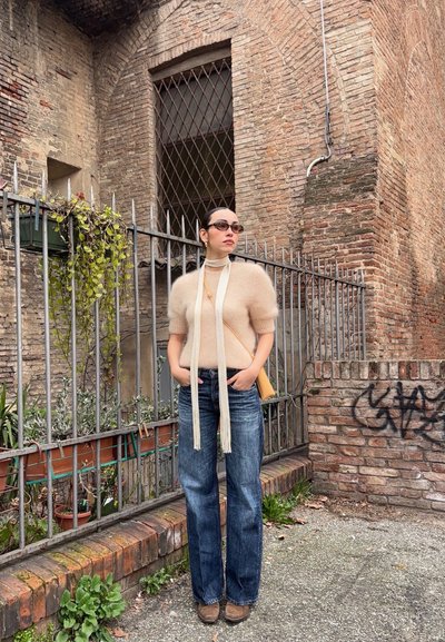 Suéter corto de color beige fuzzy, jeans oscuros acampanados y un bolso amarillo. Una bufanda blanca larga cae; unas gafas de sol negras completan el look.