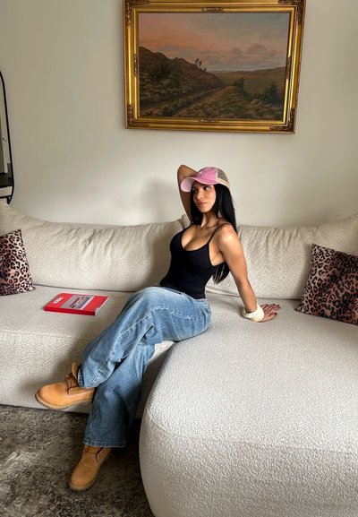 Mujer con camiseta negra sin mangas, vaqueros azules, botas color cuero claro y gorra rosa sentada en un sofá beige con cojines de estampado de leopardo y un cuadro de paisaje encima.