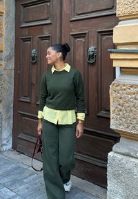 Woman smiling and standing by large wooden doors with lion head knockers, wearing a green sweater, yellow shirt, wide trousers, and white trainers.