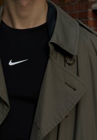 Kaki trenchcoat med stor krage og knappdetaljer, lagd over en tettsittende svart Nike t-skjorte. Glatt stoff og strukturert design.