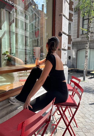 Top negro con espalda abierta, pantalones negros y zapatillas blancas. Sosteniendo una taza blanca grande, sentado en una silla plegable roja junto a una ventana.