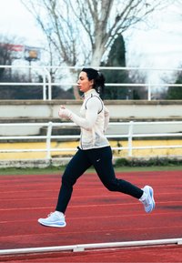 Chaussures de course bleu clair, pantalons noirs ajustés et un haut sans manches beige avec une fermeture éclair. Elle est sur une piste rouge avec des arbres en arrière-plan.
