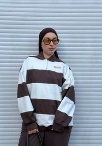 Ruskeavalkoraidallinen kauluksellinen pusero, jossa on etumainen logo. Pitää tumman käsilaukun ja käyttää erottuvia keltaisensävyisiä aurinkolaseja.