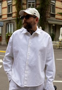 Camicia bianca a maniche lunghe con una vestibilità rilassata, apertura con bottoni e orlo curvo. Indossata con un cappellino beige chiaro e occhiali da sole.