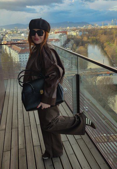 Mujer con chaqueta y pantalones de cuero marrón, boina negra, gafas de sol, sosteniendo bolso negro, de pie en un balcón con vista a la ciudad y al río.