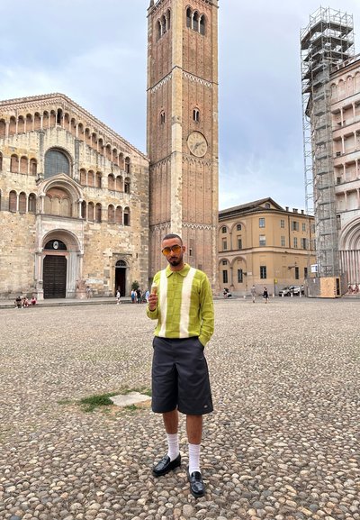 Suéter a rayas verde y crema combinado con pantalones cortos oscuros. Calcetines blancos y zapatos negros brillantes. Edificios de ladrillo y torre del reloj en el fondo.
