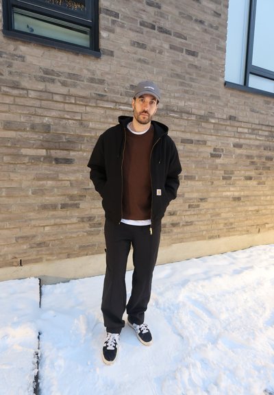 Hombre con chaqueta negra, camisa marrón, pantalones negros y gorra, de pie sobre suelo cubierto de nieve frente a una pared de ladrillo con ventanas.