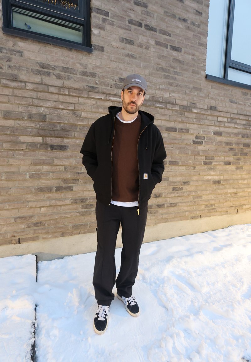 Homme portant une veste noire, une chemise brune, un pantalon noir et une casquette, debout sur un sol enneigé devant un mur de briques avec des fenêtres.