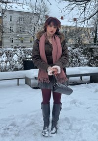 Veste matelassée marron avec capuche en fourrure, écharpe rose, sac à main noir, bottes grises montant jusqu'au genou et collants bordeaux. Arrière-plan enneigé.