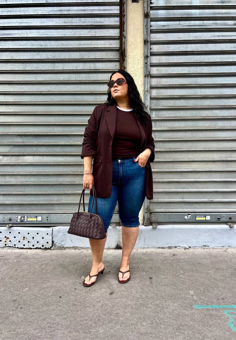 Blazer bordeaux sobre um top castanho, combinado com calções de ganga azuis. Uma bolsa tecida escura e chinelos castanhos completam o visual contra um fundo metálico.