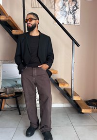 Μαύρη σακάκι πάνω από ένα καφέ εφαρμοστό μπλουζάκι, συνδυασμένο με καφέ φαρδιές παντελόνες και μαύρες μπότες. Διαθέτει πλεξούδες μαλλιά και στρογγυλά πορτοκαλί γυαλιά ηλίου.