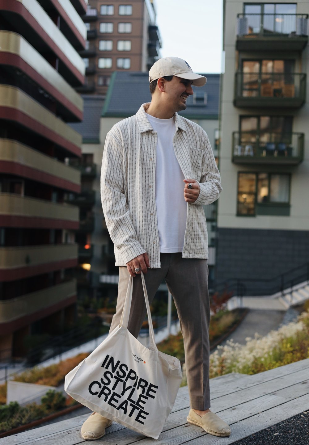 Cămașă cu dungi crem, tricou alb, pantaloni bej, pantofi slip-on bej și o geantă de pânză cu text negru. Cadru urban.