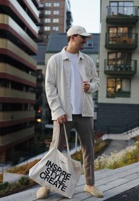 Cămașă cu dungi crem, tricou alb, pantaloni bej, pantofi slip-on bej și o geantă de pânză cu text negru. Cadru urban.