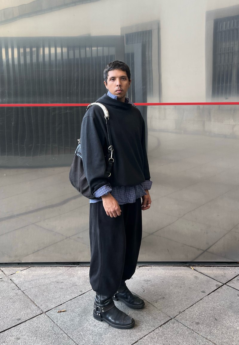 Μαύρο oversized πουλόβερ πάνω από ένα μπλε καρό πουκάμισο, χαλαρές μαύρες παντελόνες και μαύρες μπότες με ασημένια στοιχεία, στέκεται μπροστά σε ανακλαστικό φόντο.