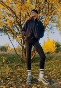 Fleecinė striukė, juodos tamprės ir raštuoti sportbačiai. Akiniai nuo saulės ir kepurė užbaigia aprangą prieš geltonų lapų foną.