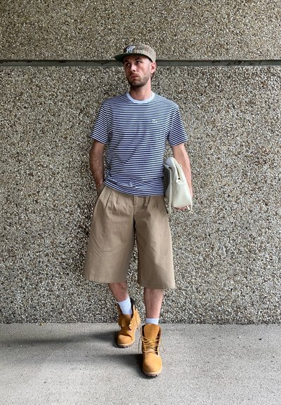 Camiseta rayas azul marino y blanca, pantalones cortos beige, botas amarillas, calcetines blancos y gorra a cuadros, sosteniendo una bolsa blanca contra una pared texturizada.