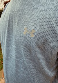 Camisola atlética azul-claro com um padrão texturizado, apresentando um pequeno logótipo prateado da Under Armour no peito. Mangas curtas e gola redonda.