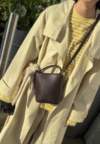 Brown leather bucket bag with a textured finish, featuring a top handle and a long strap, worn with a light yellow coat and striped shirt.