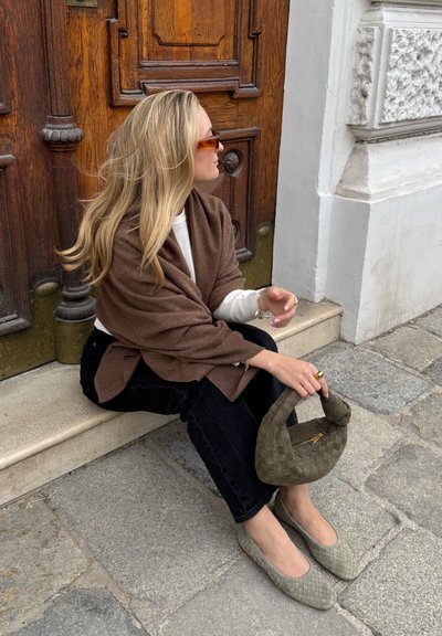 Chal marrón texturizado sobre una blusa blanca, combinado con jeans negros. Bolso tejido en color verde oliva y bailarinas a juego sobre una superficie de piedra.