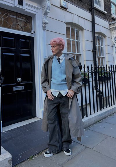 Persona con cabello rosa que lleva un abrigo gris, chaleco azul, camisa blanca, corbata negra, jeans anchos negros y zapatillas negras, de pie en una acera.