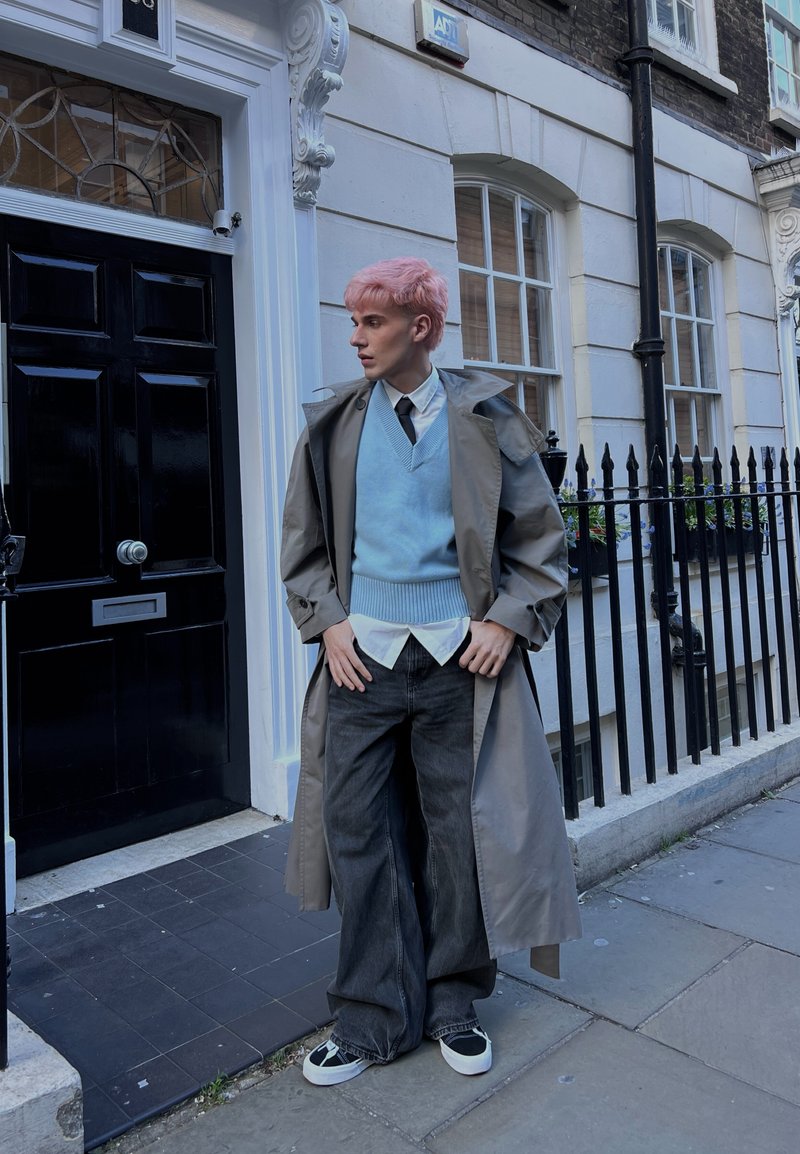 Personne aux cheveux roses portant un trench-coat gris, un gilet en laine bleu, une chemise blanche, une cravate noire, un jean noir ample et des baskets noires, debout sur un trottoir.