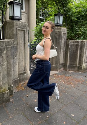 Top corto de encaje blanco, jeans acampanados de color azul oscuro y zapatillas grises, con un pequeño bolso blanco. El fondo presenta vegetación y paredes de piedra.
