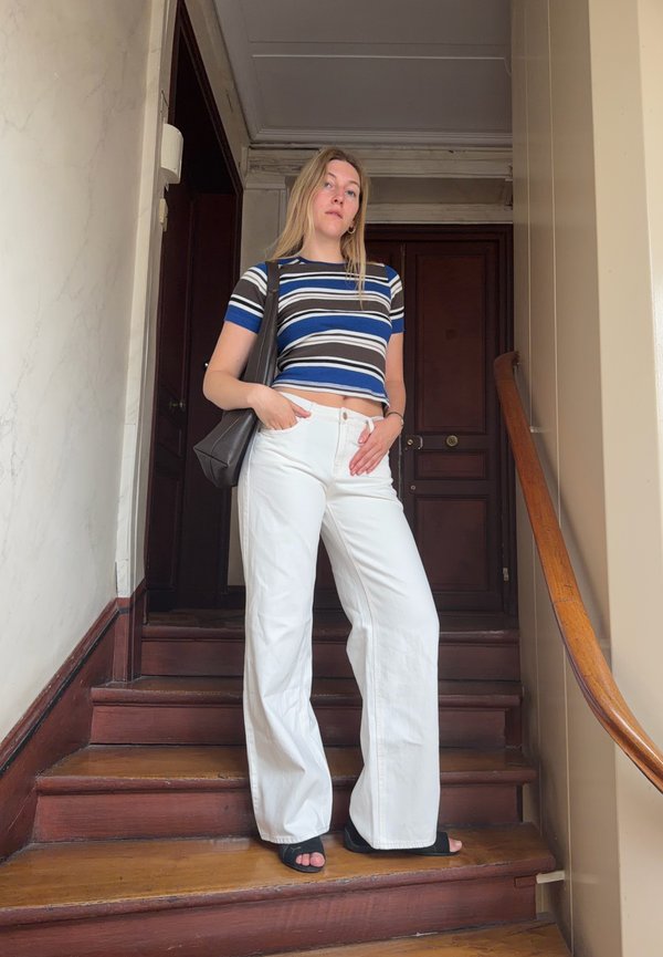 Jeune femme debout sur des escaliers en bois, portant un crop top rayé, un pantalon blanc, des sandales noires et tenant un sac à main noir.