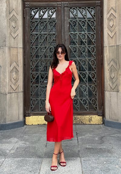 Vestido midi rojo con tirantes rizados y un detalle de lazo en la parte delantera. Combinado con tacones rojos de tiras y un pequeño bolso marrón.