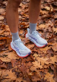 Vaaleansiniset juoksukengät, joissa on violetit yksityiskohdat ja teksturoitu verkkopäällinen. Punamusta pohja, jossa on vaimennusta. Harmaat nilkkasukat. Taustalla lehtiä.