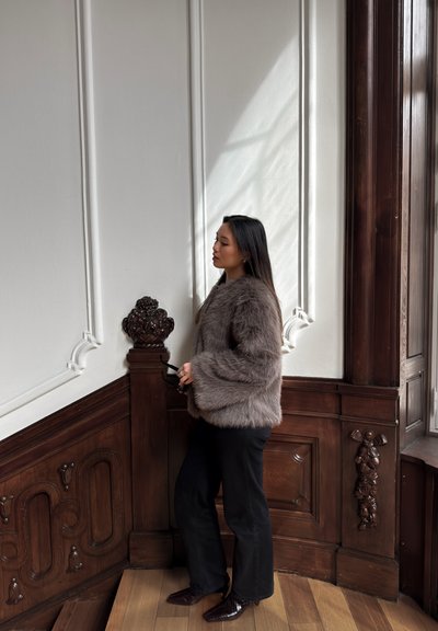 Chaqueta de pelaje sintético marrón con mangas anchas, llevada sobre pantalones negros. El entorno presenta acentos de madera y una luz suave que entra por una ventana.