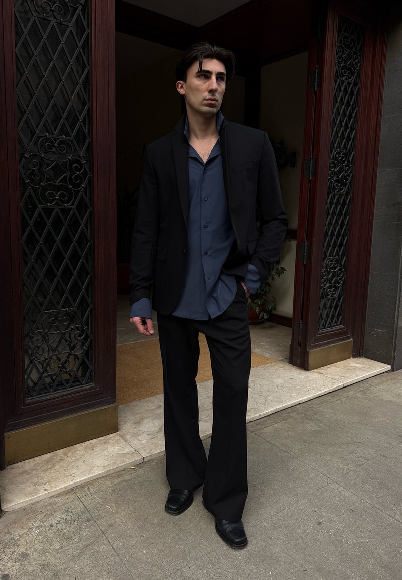 Homme en costume noir et chemise bleue, debout à une porte avec des panneaux en fer décoratifs, regardant sur le côté, une main dans la poche.