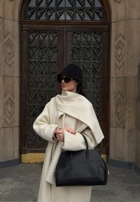 Črna usnjena torbica z zvitimi ročaji, združena s belo volna ruto in črno pleteno kapo; stoji ob okrašenih kovinskih vratih.